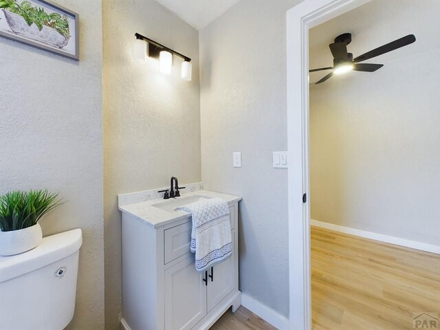 half bath with toilet, ceiling fan, vanity, wood finished floors, and baseboards