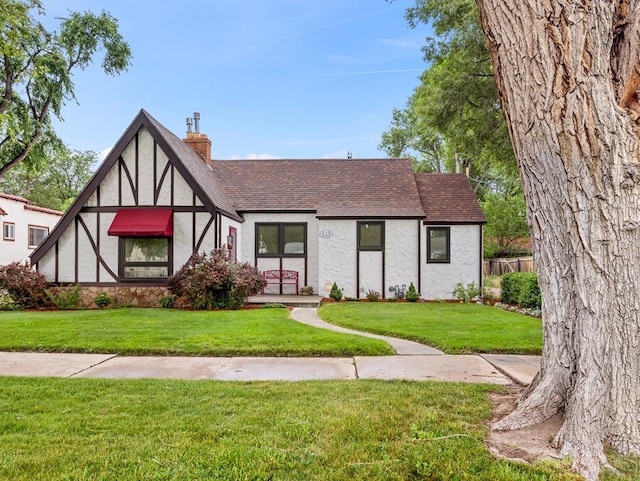 tudor house featuring a front lawn, stucco siding, roof with shingles, and a chimney