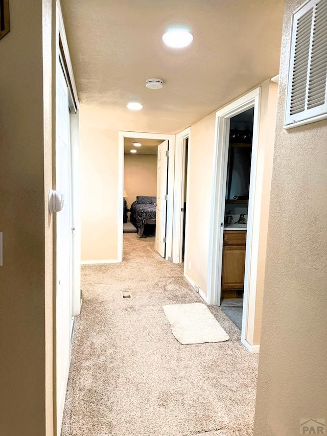 hallway featuring a textured wall, visible vents, baseboards, and light colored carpet