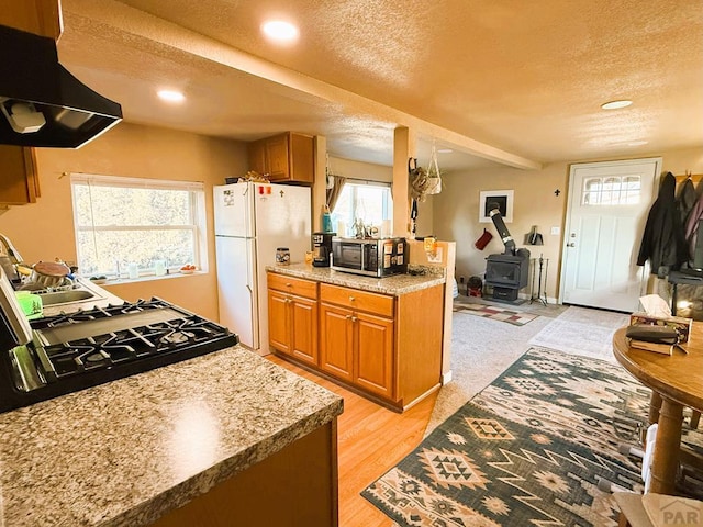 kitchen featuring brown cabinets, stainless steel microwave, freestanding refrigerator, a wood stove, and exhaust hood