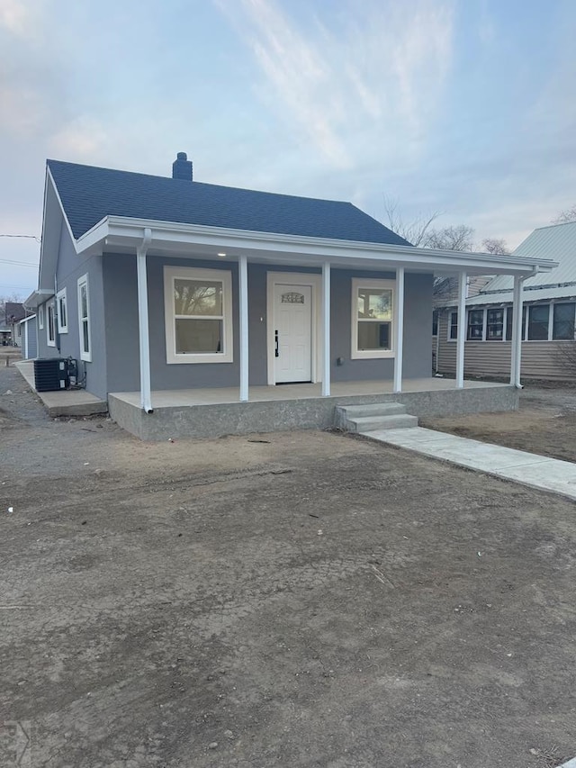 view of front of home with a shingled roof, a chimney, a porch, central AC, and stucco siding