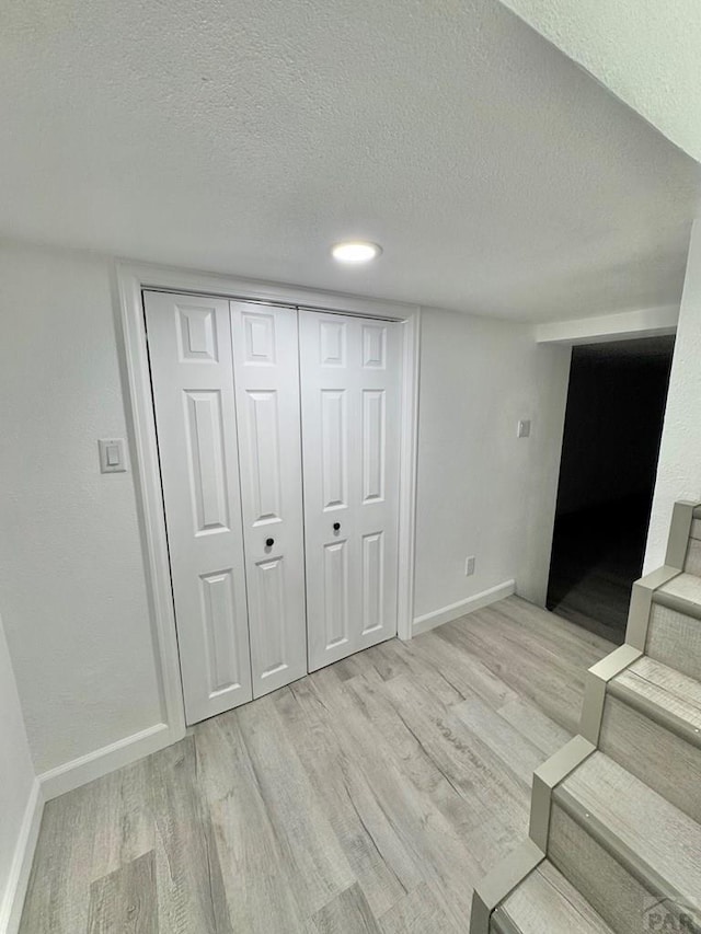 basement with a textured ceiling, stairway, light wood-style flooring, and baseboards