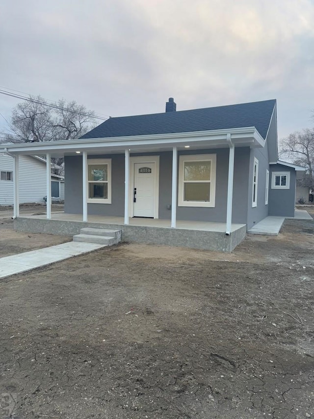 bungalow-style home with covered porch, roof with shingles, a chimney, and stucco siding