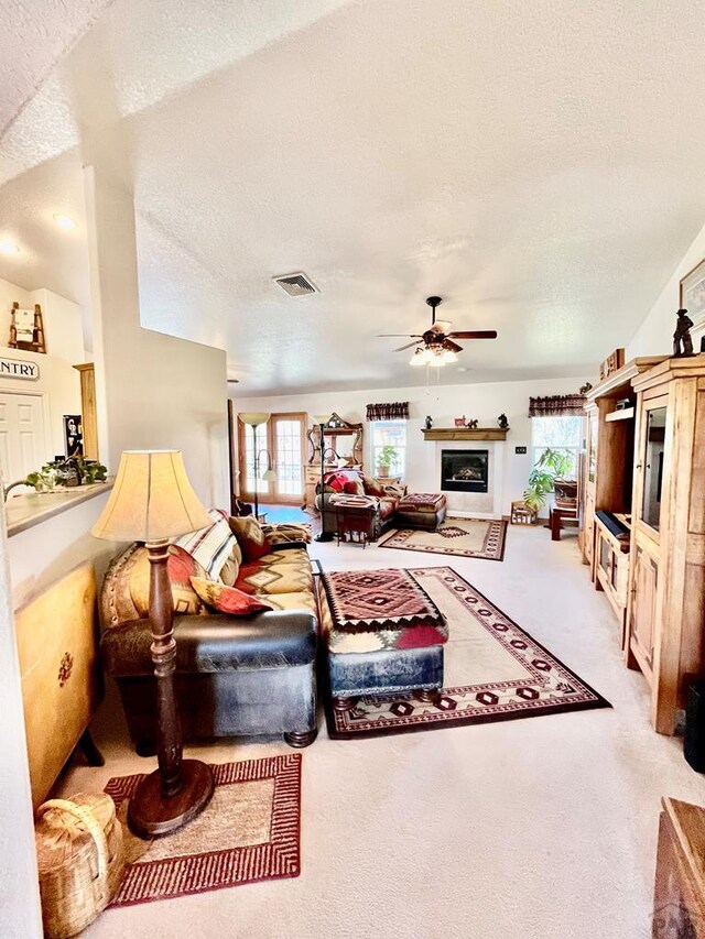 living room featuring visible vents, a glass covered fireplace, light colored carpet, ceiling fan, and a textured ceiling