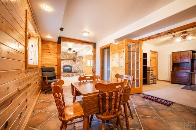 dining area featuring a ceiling fan, dark colored carpet, a stone fireplace, wood walls, and dark tile patterned floors