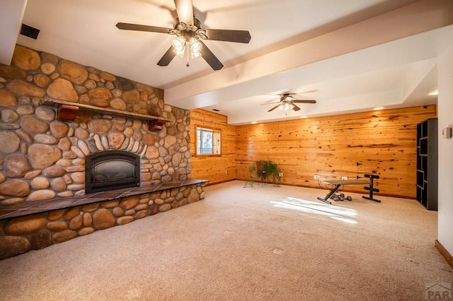 unfurnished living room with carpet, beam ceiling, ceiling fan, wooden walls, and a stone fireplace