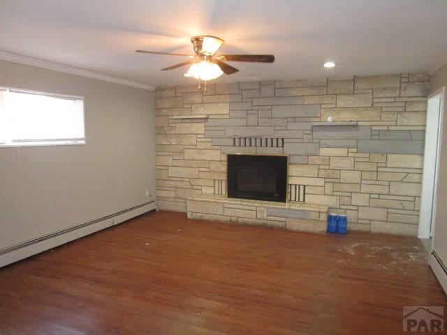 unfurnished living room featuring a ceiling fan, dark wood-style floors, a baseboard radiator, ornamental molding, and a stone fireplace