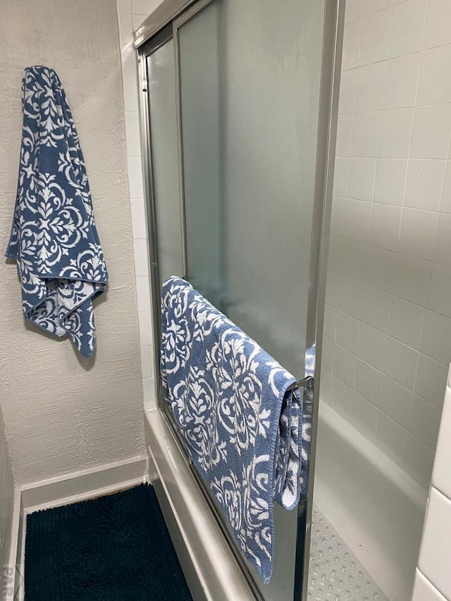 full bath with combined bath / shower with glass door, baseboards, and a textured wall
