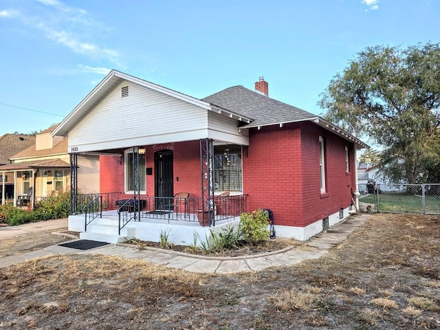 bungalow-style home with brick siding, a chimney, a shingled roof, covered porch, and fence