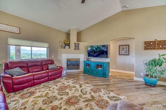 living room with ceiling fan, high vaulted ceiling, baseboards, light wood finished floors, and a tiled fireplace