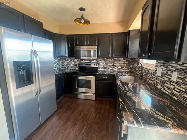 kitchen with stainless steel appliances, backsplash, dark wood-type flooring, a sink, and dark stone counters