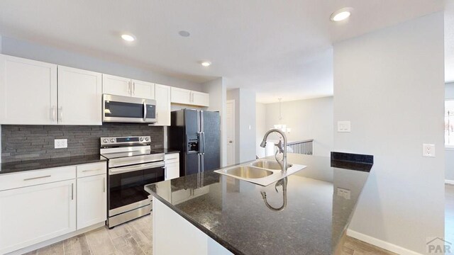 kitchen with a center island with sink, stainless steel appliances, white cabinets, a sink, and dark stone counters