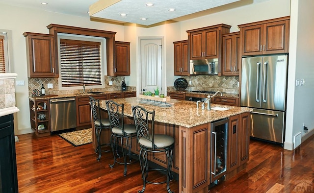 kitchen with light stone counters, a center island with sink, appliances with stainless steel finishes, dark wood-type flooring, and beverage cooler