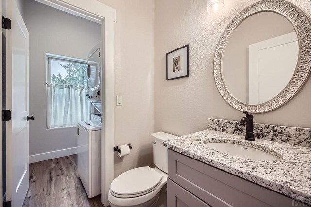 half bath featuring stacked washer and dryer, toilet, vanity, wood finished floors, and baseboards