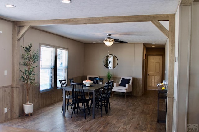 dining room featuring a ceiling fan, a wainscoted wall, dark wood-type flooring, a textured ceiling, and beam ceiling