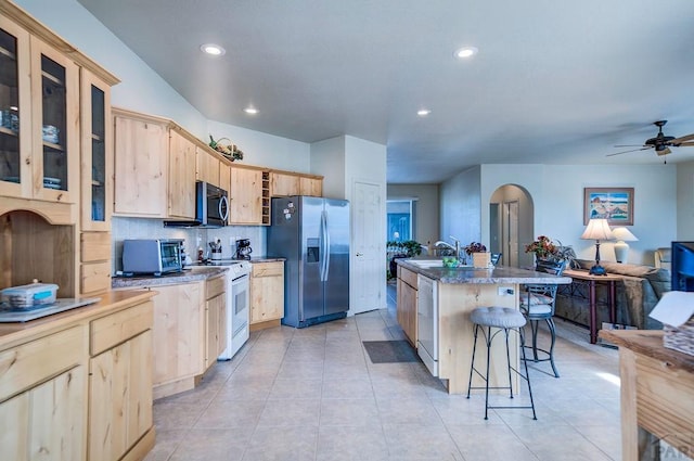 kitchen with arched walkways, appliances with stainless steel finishes, light brown cabinetry, an island with sink, and glass insert cabinets