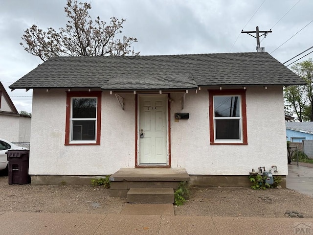 bungalow featuring stucco siding, roof with shingles, and fence