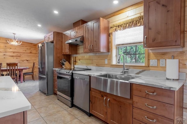 kitchen with wooden walls, stainless steel appliances, under cabinet range hood, pendant lighting, and a sink