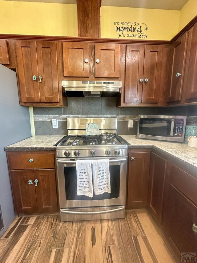 kitchen with light stone counters, under cabinet range hood, appliances with stainless steel finishes, light wood-type flooring, and decorative backsplash