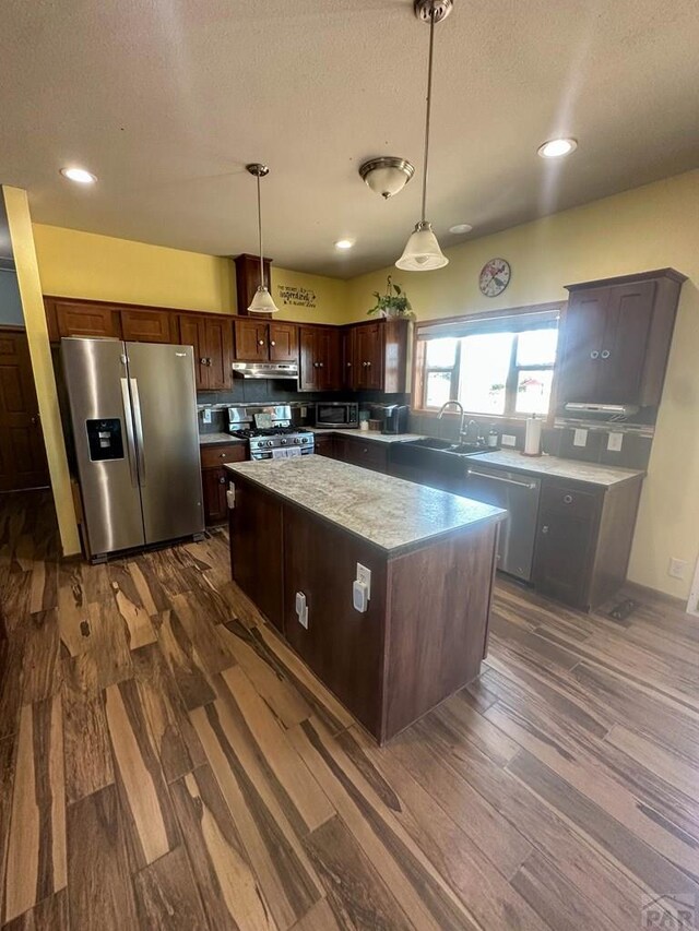 kitchen with stainless steel appliances, a sink, a kitchen island, dark wood finished floors, and decorative light fixtures