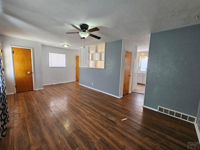 empty room with a textured wall, a ceiling fan, visible vents, baseboards, and dark wood finished floors