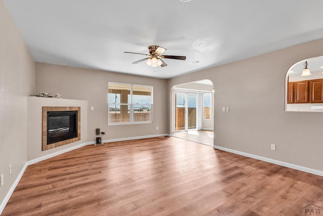 unfurnished living room with baseboards, arched walkways, a ceiling fan, a tile fireplace, and light wood-style floors