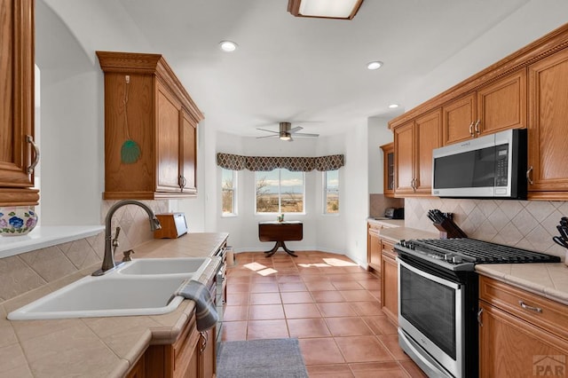 kitchen with stainless steel appliances, tile counters, brown cabinetry, light tile patterned flooring, and a sink