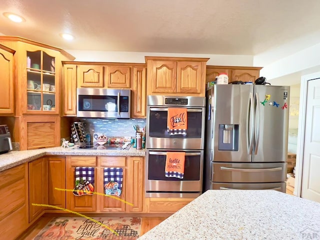 kitchen with light stone counters, stainless steel appliances, brown cabinets, tasteful backsplash, and glass insert cabinets