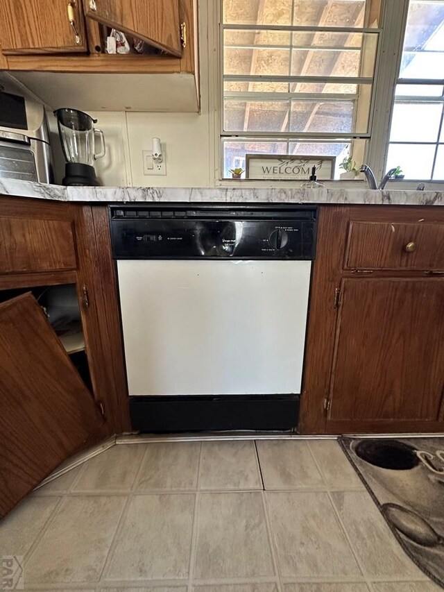 kitchen with light tile patterned floors, dishwashing machine, brown cabinetry, and light countertops