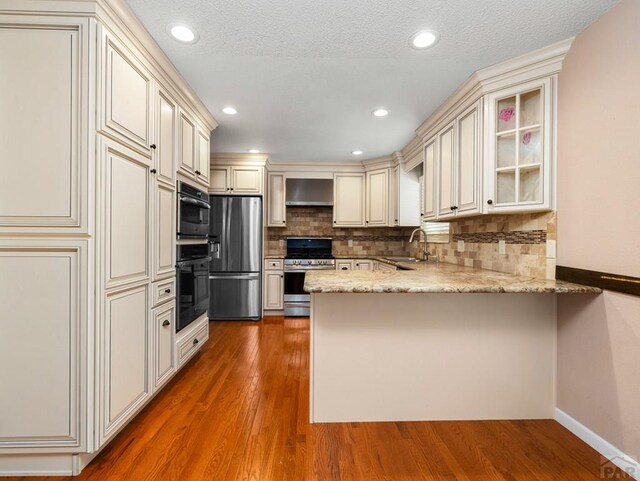 kitchen featuring glass insert cabinets, appliances with stainless steel finishes, a sink, a peninsula, and wall chimney exhaust hood