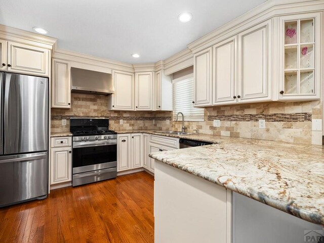 kitchen featuring light stone counters, appliances with stainless steel finishes, glass insert cabinets, a sink, and wall chimney exhaust hood