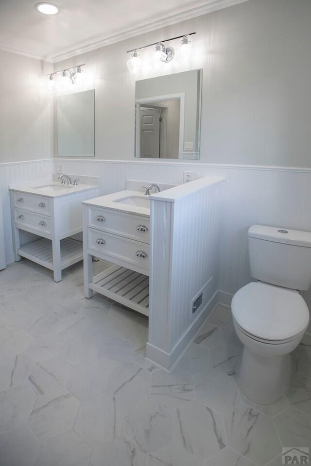 bathroom featuring marble finish floor, wainscoting, two vanities, and crown molding