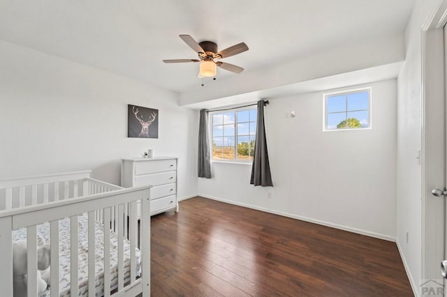 bedroom with a crib, dark wood finished floors, a ceiling fan, and baseboards