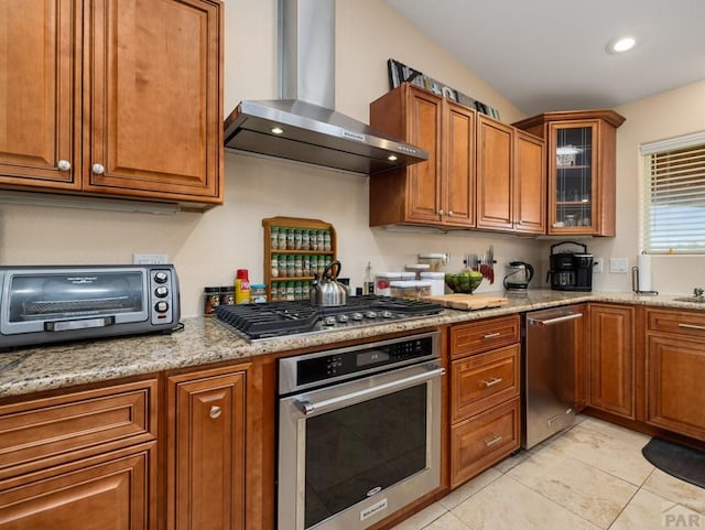 kitchen with brown cabinets, stainless steel appliances, glass insert cabinets, light stone countertops, and wall chimney exhaust hood