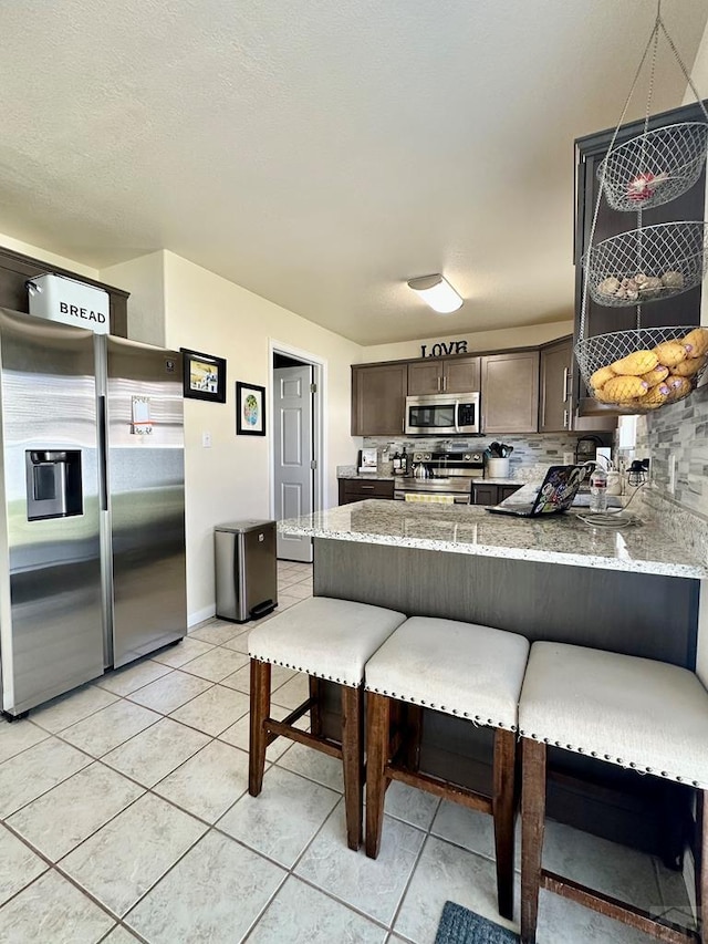 kitchen featuring a peninsula, light tile patterned flooring, light stone countertops, dark brown cabinets, and stainless steel appliances