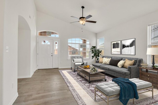 living room with ceiling fan, high vaulted ceiling, arched walkways, baseboards, and light wood finished floors