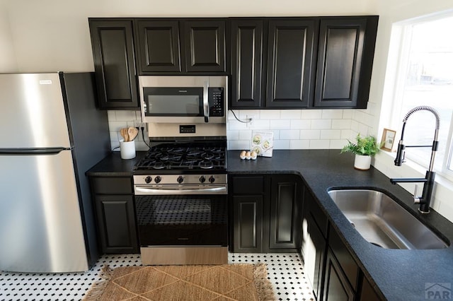 kitchen with dark cabinets, stainless steel appliances, a sink, backsplash, and dark countertops