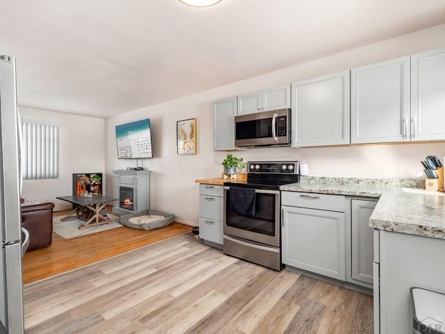 kitchen with appliances with stainless steel finishes, a lit fireplace, gray cabinets, light countertops, and light wood-type flooring