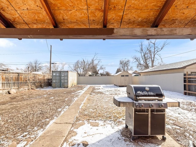 snow covered patio featuring an outbuilding, a storage unit, a fenced backyard, and a grill