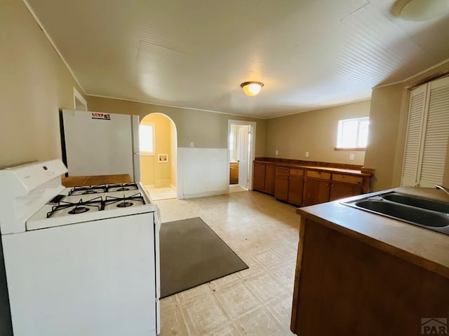 kitchen featuring arched walkways, white appliances, a sink, light floors, and brown cabinetry