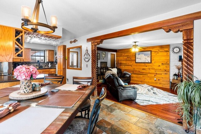 dining space featuring stone finish flooring, wooden walls, decorative columns, and ceiling fan with notable chandelier