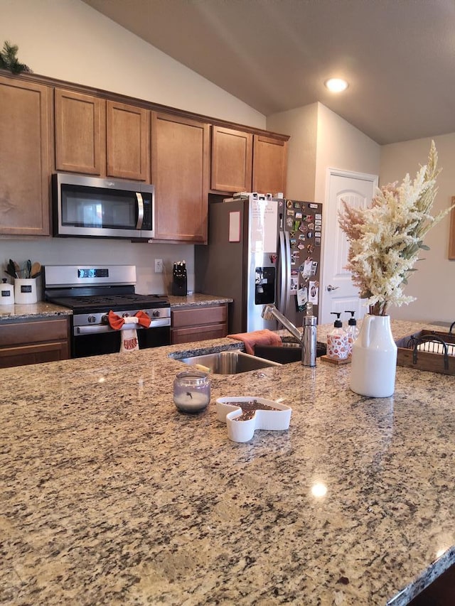 kitchen with brown cabinetry, light stone counters, vaulted ceiling, stainless steel appliances, and a sink