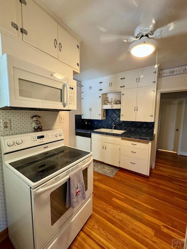 kitchen with white cabinets, a sink, dark countertops, white appliances, and light wood-style flooring
