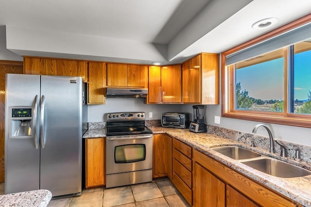 kitchen with brown cabinetry, light stone counters, appliances with stainless steel finishes, under cabinet range hood, and a sink