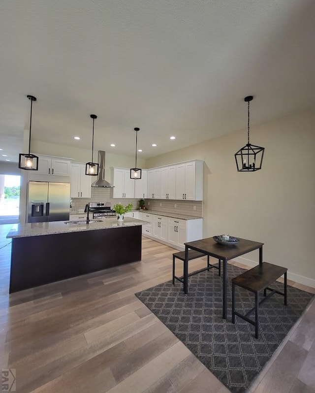 kitchen with stainless steel appliances, white cabinetry, wall chimney range hood, an island with sink, and pendant lighting