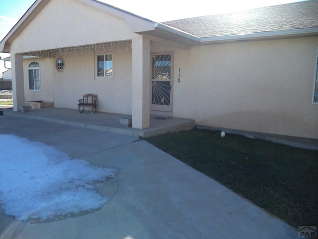 view of exterior entry with a patio, a shingled roof, and stucco siding