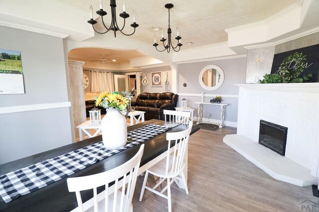 dining room featuring arched walkways, crown molding, a glass covered fireplace, wood finished floors, and ceiling fan with notable chandelier