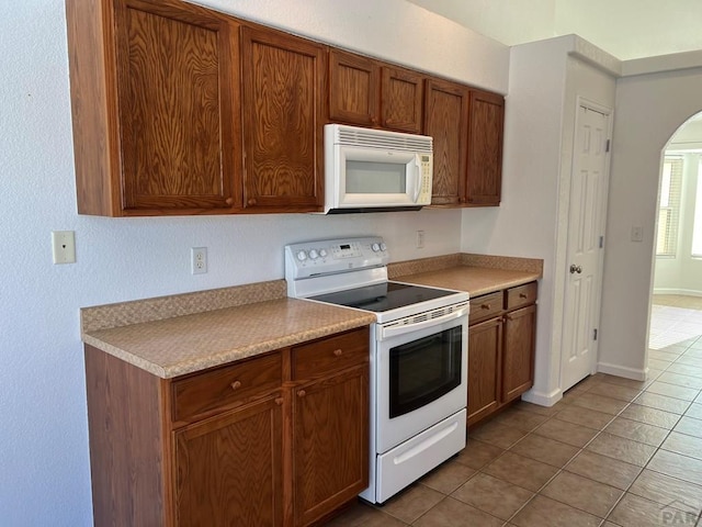 kitchen with arched walkways, light countertops, brown cabinetry, light tile patterned flooring, and white appliances