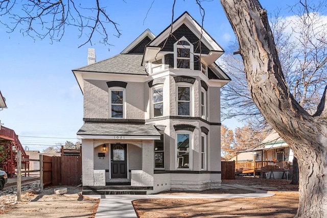 victorian house featuring roof with shingles, brick siding, a chimney, and fence