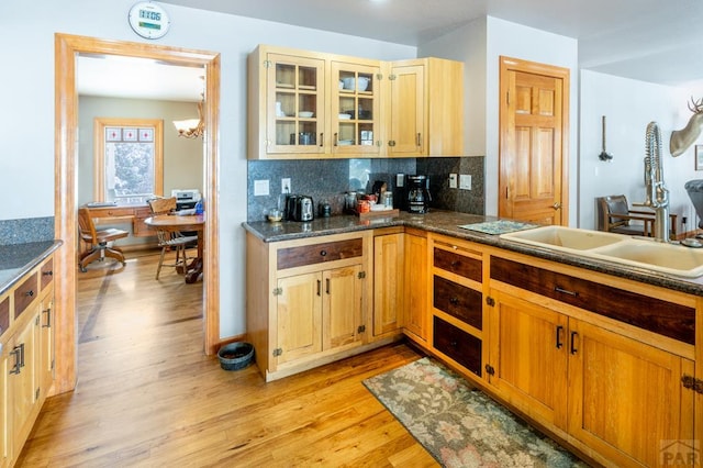 kitchen with dark countertops, glass insert cabinets, a sink, light wood-style floors, and backsplash